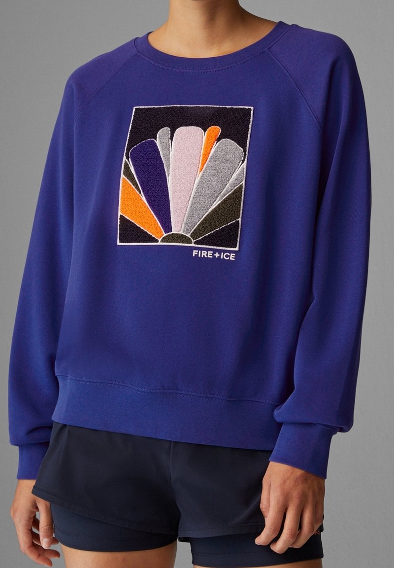 Personne portant un sweat bleu avec un patch abstrait multicolore et le texte "FIRE + ICE", associé à un short de sport noir.