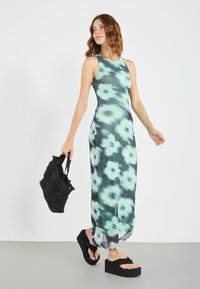 Maxikleid mit grünem Blumenmuster, das ein eng anliegendes Tank-Design aufweist. Kombiniert mit einer schwarzen Tasche und Plateau-Sandalen mit weichem Riemen.