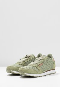 Paire de baskets en maille vert clair et daim avec des lacets beige, des accents marron et des semelles blanches tachetées sur une surface blanche.