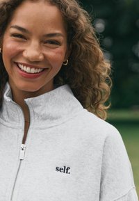 Svetle šedá mikina na zip s vysokým límcem, s malým černým vyšitým textem "self." na levé straně. Měkká, strukturovaná tkanina.