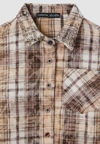 Camicia a quadri marrone e crema con colletto a polo, decorata con borchie trasparenti sulle spalle e una tasca sul petto. Tessuto in misto cotone.