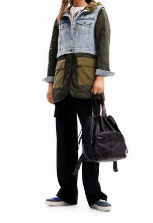 Person trägt grüne gesteppte Jacke mit Jeansweste darüber, schwarze Hose, blaue Turnschuhe, hält schwarzen Kordelzug-Rucksack mit geprägtem Muster.