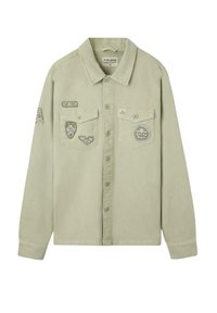 Camisa de botón color khaki claro con mangas largas, dos bolsillos en el pecho y parches y insignias de estilo militar en la parte delantera y en la manga izquierda.