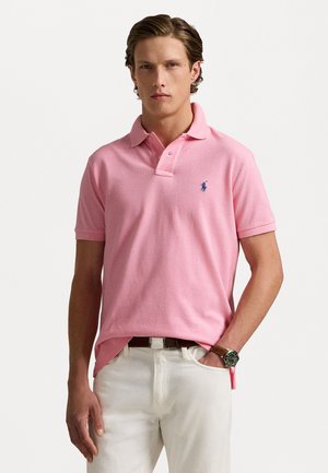 Rosa polo skjorte med krage, korte ermer og en liten blå brodert logo på brystet, parret med hvite bukser.