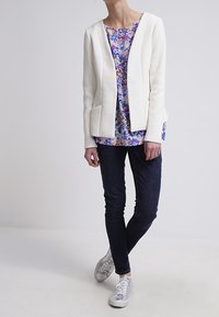 Witte textuur blazer met twee vo pockets, gelaagd over een meerkleurige bloemetjestop, gecombineerd met donkere jeans en zilveren sneakers.