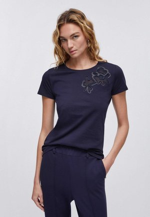 Donna con capelli biondi ondulati, che indossa una maglietta blu navy a maniche corte con dettagli ricamati a fiori sulla spalla sinistra e pantaloni abbinati.