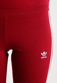Röda träningstights med en åtsittande passform, en vit Adidas-logotyp på vänster ben och vita sidostripes för extra detalj.