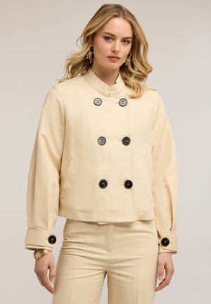 Kobieta o blond włosach, ubrana w kremową marynarkę dwurzędową z czarnymi guzikami i dopasowane spodnie, stojąca na neutralnym tle.