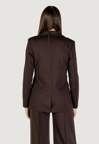 Blazer marrón a medida con mangas largas, cuello con muesca y costura trasera recta. Textura de tejido suave, combinado con pantalones de pierna ancha.