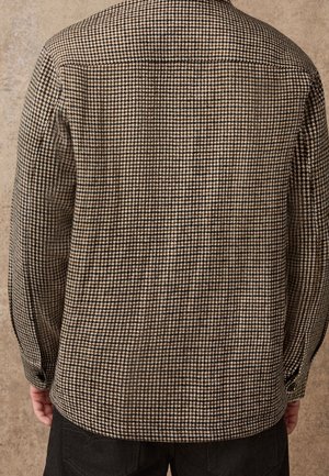 Personne portant une veste à motif pied-de-poule marron et noir, vue de dos, les mains reposant le long du corps.