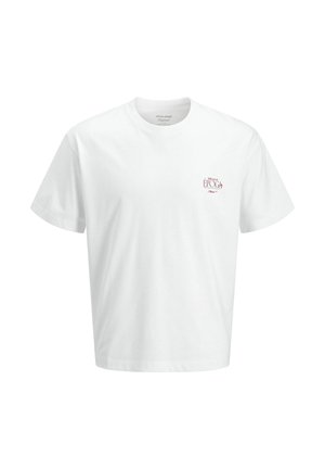 Garçon souriant portant un t-shirt blanc, un jean large foncé, des chaussures marron et une casquette rouge, debout devant un fond blanc uni.
