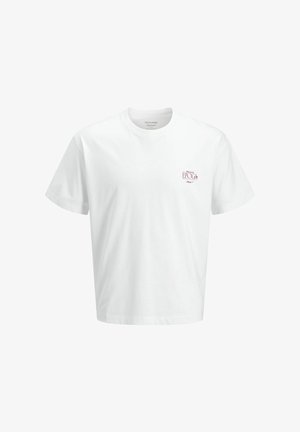Garçon souriant portant un t-shirt blanc, un jean large foncé, des chaussures marron et une casquette rouge, debout devant un fond blanc uni.