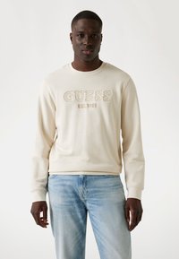 Sudadera crema con bordado "GUESS EST.1981" en un color contrastante. Mangas largas, cuello redondo, combinada con jeans azul claro.