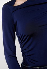 Close-up van een persoon die een marineblauw langarmtop draagt met geplooide stofdetails aan de zijkant en donkerblauwe jeans.