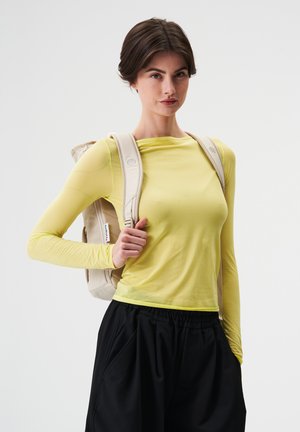 Young woman wearing a yellow long-sleeve top, black pants, and a beige backpack, standing against a plain white background.
