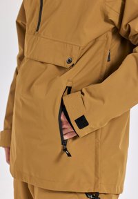 Heren bruine outdoorjack, waterdichte stof, met een voorzak en zijzak met rits, voorzien van zwarte accenten en klittenbandsluiting bij de manchetten.