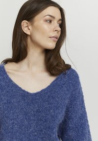 Pull en tricot bleu avec un col en V. Le tissu semble doux et flou, avec une coupe ample et une finition texturée. Design minimaliste, sans ornements.