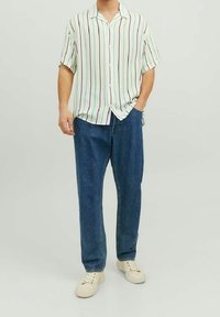 Mężczyzna w białej koszuli z krótkim rękawem w pionowe zielone i czarne paski, w niebieskich dżinsach i białych trampkach, stoi z jedną ręką w kieszeni.