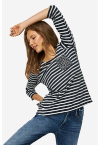 Maglietta a righe navy e bianche con maniche a 3/4, comodamente dotata di una tasca a pois. Il tessuto sembra morbido e ha una vestibilità casual. Indossata con jeans blu.
