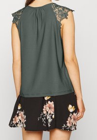 Ärmlös mörkgrön topp med spetsiga blommönster på axlarna, i kombination med en svart kjol med blommönster och plisserad design.