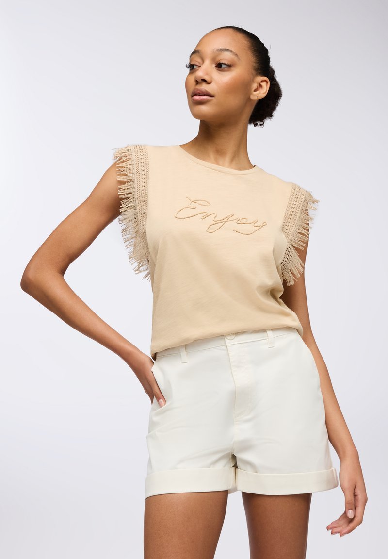 Top in cotone beige senza maniche con dettagli frangiati alle maniche e ricamo "Enjoy". Abbinato a pantaloni corti bianchi con risvolto. Texture morbida.