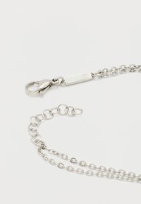 Ασημένιο κολιέ αλυσίδα με κούμπωμα καβουριού και ορθογώνια ταμπέλα χαραγμένη με "LACOSTE" σε λευκό φόντο.