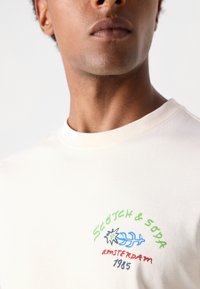 Krämfärgad bomulls t-shirt med rund halsringning, som har en färgglad broderad logotyp med grön text och en blå växtdesign.