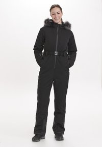 Svart isolerad jumpsuit med fuskpälshette, bälte i midjan, dragkedja fram och fickor. Slät textur med en stilren, modern design.