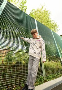 Jasno beżowy hoodie z czarnymi wzorami w formie doodli, w połączeniu z szarymi szerokimi spodniami oraz czarnymi sneakersami, na tle zielonego metalowego ogrodzenia.