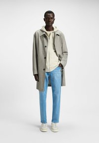 Beige ternet lang frakke, hvid hoodie under, lyseblå jeans og hvide sneakers. Glat stof med synlige knapper og lommer.