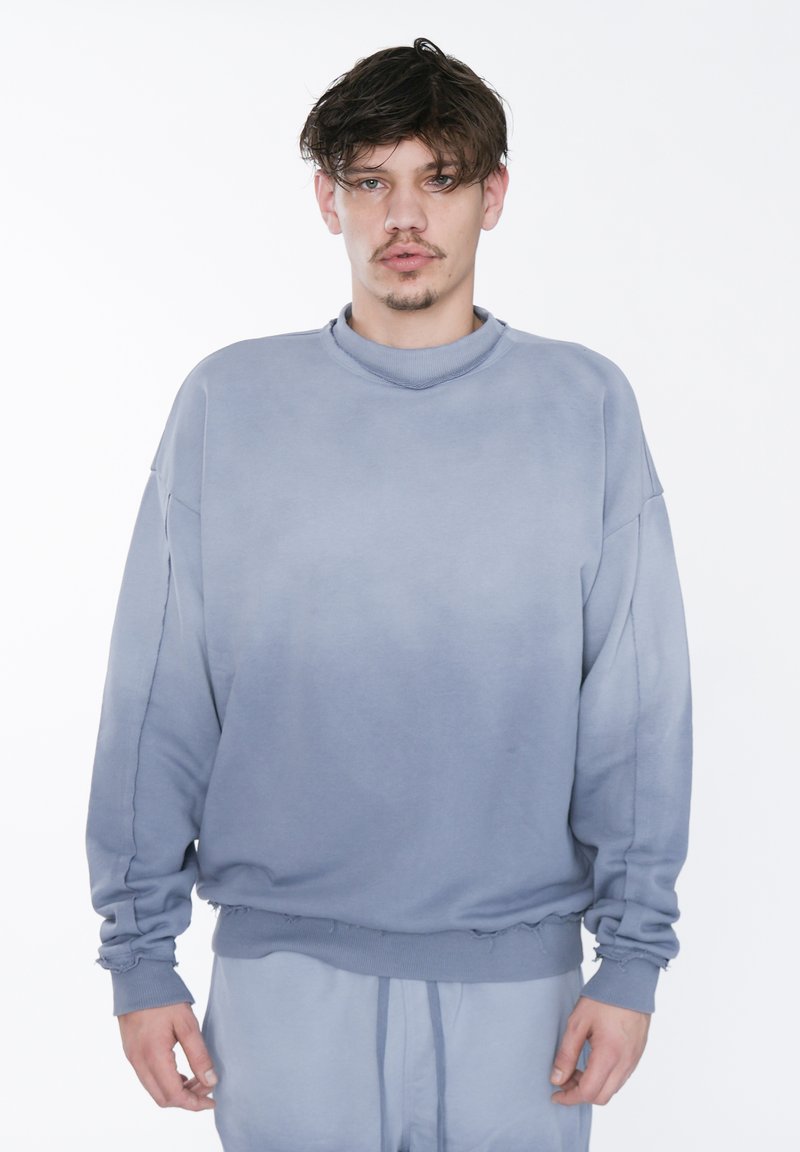 Sudadera azul claro con un cuello alto, hombros caídos y dobladillo deshilachado, hecha de material suave; presenta una textura en degradado.
