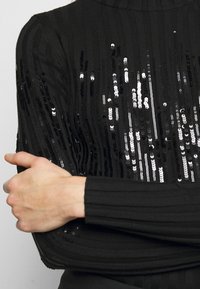 Suéter negro de canalé con filas verticales de lentejuelas plateadas y negras alternas en la parte frontal. Manga larga y diseño de cuello alto.