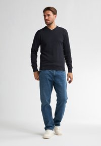 Navyblauer V-Ausschnitt-Pullover aus glattem Strickmaterial, kombiniert mit blauen Jeans. Das Modell steht lässig da und trägt weiße Sneaker.