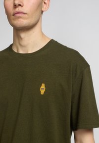 Camiseta de algodón verde oscuro con mangas cortas y escote redondo. Cuenta con un pequeño gráfico de un reloj amarillo bordado en el lado izquierdo del pecho.