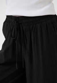 Personne portant un pantalon noir ample à taille élastique avec cordon, associé à un haut blanc côtelé.