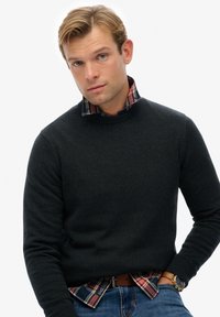 Jeune homme aux cheveux blonds portant un pull noir à col ras du cou, une chemise à carreaux en dessous, un jean bleu, une ceinture marron et une montre-bracelet en or.