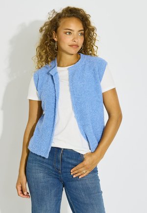 Mujer con pelo rizado que lleva pendientes de aro, una camiseta blanca, un chaleco texturizado sin mangas de color azul claro y vaqueros azules, de pie frente a un fondo blanco.