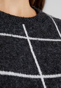 Gros plan d’un pull noir duveteux avec des lignes blanches à carreaux porté par une personne montrant l’encolure et une partie du cou.