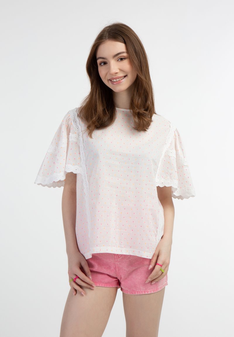 Blusa bianca a maniche corte con pois rosa, bordo in pizzo sulle spalle, abbinata a pantaloni corti rosa chiaro. Texture liscia, design casual.