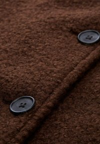 Bruine textuur stof met een fluweelachtige uitstraling, voorzien van twee zwarte knopen die op het oppervlak zijn genaaid, wat zorgt voor een kleur- en afwerkingscontrast.