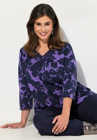 Lila V-kauluspaita mustalla kukkakuviolla, kolmannes hihat ja sileä kangas; yhdistetty tummiin housuihin ja metallisiin jalkineisiin.