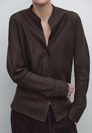 Personne portant une chemise marron foncé à manches longues avec col mandarin, un bras plié sur le torse, sur fond uni.