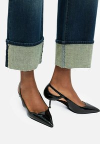 Tacones slingback de charol negro con punta afilada y un pequeño tacón de aguja. Llevados con jeans de mezclilla oscuros que presentan puños de mezclilla clara.