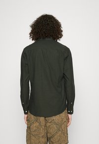 Cămașă cu mâneci lungi de un verde închis, cu colorat standard și manșete cu nasturi, fabricată dintr-un material texturat. Detalii vizibile de cusătură pe spate.