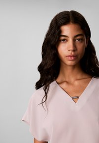 Jeune femme aux longs cheveux bouclés et foncés, portant un haut rose clair à col en V sur un fond gris uni.