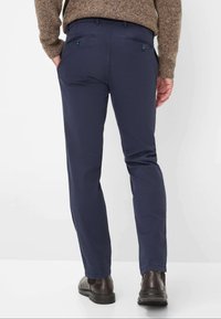 Marineblaue Chinos mit schmaler Passform, ausgestattet mit Gesäßtaschen mit Knopfverschluss und einem glatten, nicht strukturierten Stoff. Kombiniert mit dunkelbraunen Schuhen.