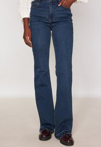 Persona che indossa jeans a vita alta di colore blu scuro con orli sfrangiati e mocassini borborigmi bordeaux, mano appoggiata sull'anca.