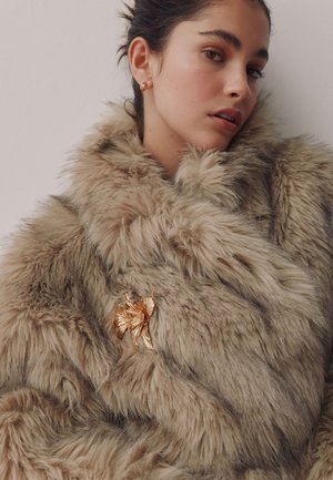 Woman wearing a tan faux fur coat with a gold floral brooch pinned on the chest, looking slightly to the side against a plain background.