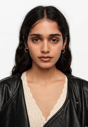 Joven mujer con cabello largo y oscuro lleva pendientes de aro con diseño floral, una blusa blanca de encaje y una chaqueta de cuero negra, mirando directamente hacia adelante.