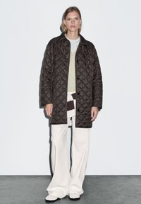 Prošívaná hnědá bunda s knoflíkovým zapínáním, nošená přes světle zelený top a krémové kalhoty s širokými nohavicemi, s různými texturami.